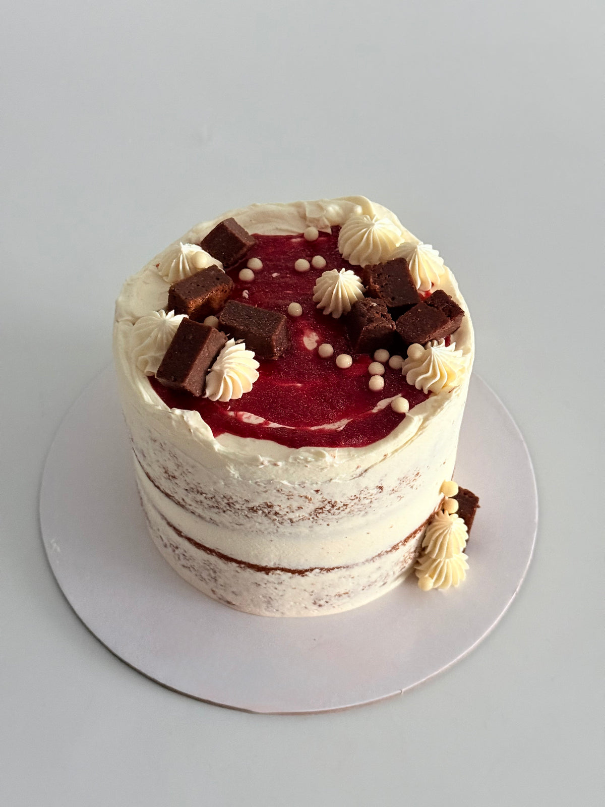 A semi-naked cake with white frosting, a red fruit topping, brownie chunks, and piped cream decorations, presented on a white round board