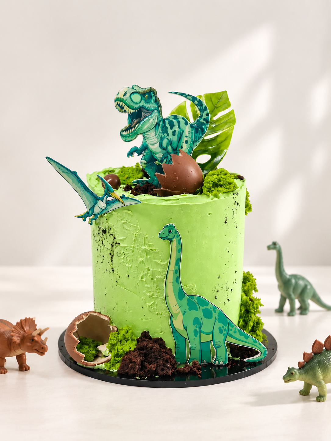 A green frosted birthday cake decorated with dinosaur figurines, a chocolate egg, faux moss, and a chocolate leaf, displayed on a white surface with small toy dinosaurs around it.