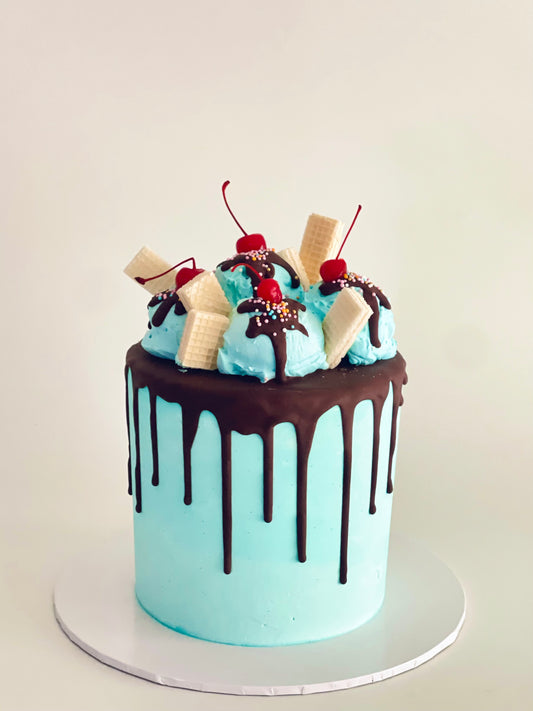 layered cake with chocolate drip, scoops of buttercream to look like ice cream sundae, topped with cherries, wafers, chocolate sauce and sprinkles