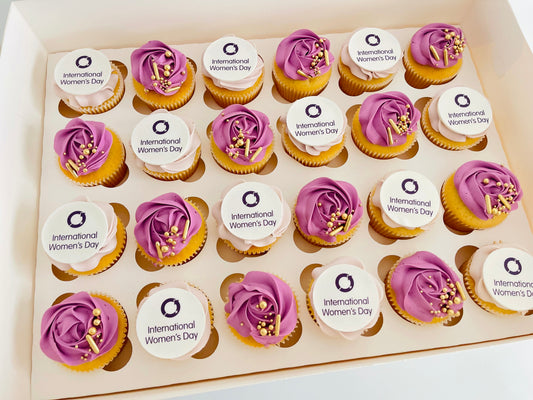 A box of cupcakes decorated with International Women's Day themed toppers and purple sprinkles.